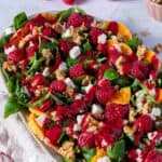 Das Bild zeigt eine ansprechende Aufnahme eines farbenfrohen Wintersalats, ideal als festliche Vorspeise für die Weihnachtszeit. Auf einem länglichen, ovalen Teller ist ein üppiger Feldsalat mit Rucola angerichtet, gespickt mit leuchtend roten Himbeeren, saftigen Orangenfilets, weißen Feta-Würfeln und knackigen Walnüssen. Ein kräftig rotes Himbeerdressing überzieht den gesamten Salat und verleiht ihm eine elegante, glänzende Note.