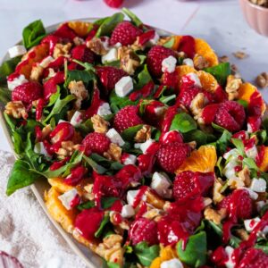 Das Bild zeigt eine ansprechende Aufnahme eines farbenfrohen Wintersalats, ideal als festliche Vorspeise für die Weihnachtszeit. Auf einem länglichen, ovalen Teller ist ein üppiger Feldsalat mit Rucola angerichtet, gespickt mit leuchtend roten Himbeeren, saftigen Orangenfilets, weißen Feta-Würfeln und knackigen Walnüssen. Ein kräftig rotes Himbeerdressing überzieht den gesamten Salat und verleiht ihm eine elegante, glänzende Note.