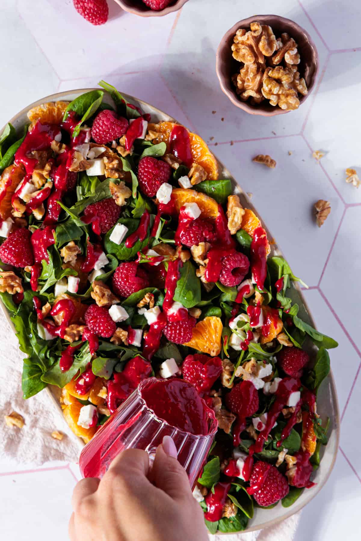 Eine große, ovale Schale, gefüllt mit Feldsalat, Rucola, Orangenfilets, Himbeeren, Fetawürfeln und Walnüssen, steht auf einer hellen, gefliesten Oberfläche. Eine Hand ist dabei zu sehen, wie sie ein Glas mit leuchtend rotem Himbeerdressing über den Salat hält und das Dressing über die Zutaten gießt. Im oberen Teil des Bildes sind zwei kleine Schalen sichtbar, eine mit Himbeeren und die andere mit Walnüssen. Das Foto ist von oben aufgenommen und zeigt eine gut ausgeleuchtete Szene.