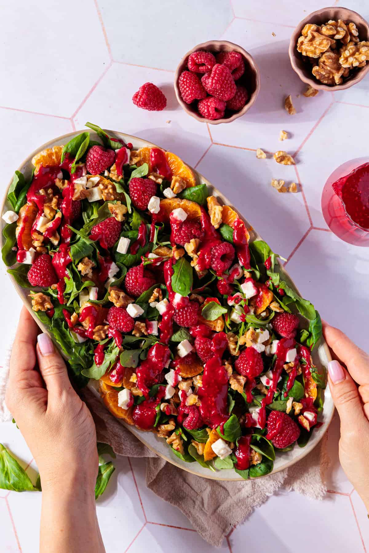 Ein großformatiges Foto zeigt einen frischen Salat, der auf einem ovalen, cremefarbenen Teller angerichtet ist. Der Salat besteht aus Feldsalat, Rucola, orangefarbenen Orangenfilets, weißen Fetawürfeln, ganzen roten Himbeeren und grob gehackten Walnüssen. Über den Salat ist ein leuchtend rotes, cremiges Himbeerdressing geträufelt. Der Teller wird von zwei Händen gehalten, die ihn von den Seiten in die Mitte des Bildes schieben. Im oberen Teil des Bildes sind zwei kleine Schalen zu sehen: eine enthält zusätzliche Himbeeren, die andere Walnüsse. Ein Glas mit dem Himbeerdressing steht rechts daneben. Der Hintergrund ist eine helle, geflieste Arbeitsfläche.