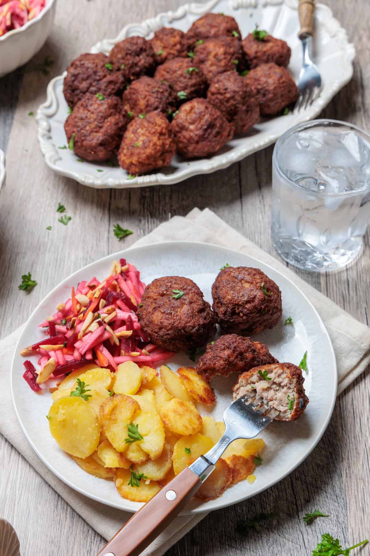 Angerichtete Portion von Thermomix®-Frikadellen auf einem Teller, serviert mit knusprigen Bratkartoffeln und einem bunten Rote-Bete-Salat. Eine angeschnittene Frikadelle zeigt die saftige Textur des Hackfleisch-Gerichts, das mit frischer Petersilie garniert ist. Im Hintergrund ist eine weitere Platte mit Frikadellen und ein Wasserglas zu sehen.