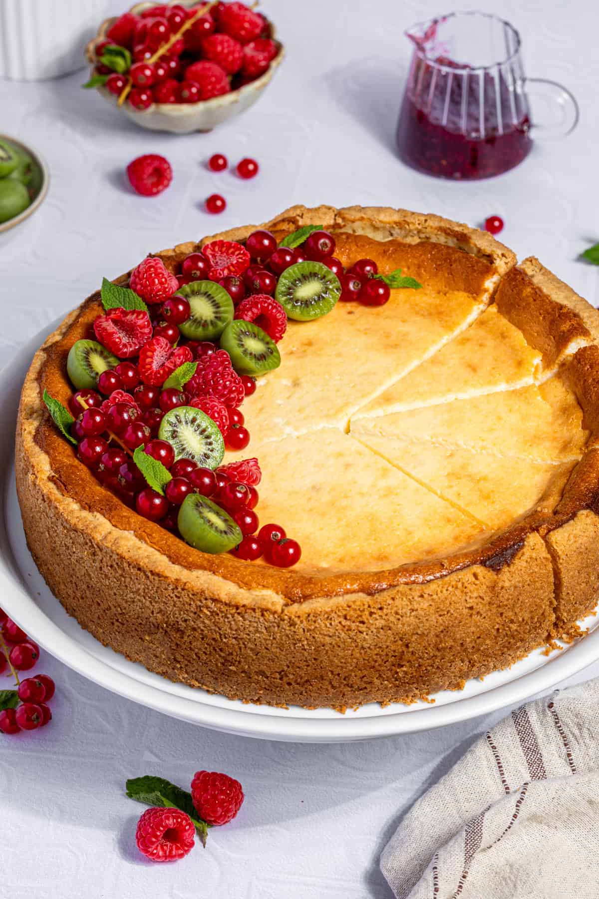 Ein ganzer, leicht angeschnittener Käsekuchen mit Himbeerboden auf einer weißen Tortenplatte. Der cremige Cheesecake ist auf der golden gebackenen Oberfläche reichlich mit frischen, roten Himbeeren, glänzenden roten Johannisbeeren und kleinen, grünen Mini-Kiwi-Beeren dekoriert. Einige Kuchenstücke sind bereits abgetrennt und zeigen die homogene, helle Füllung und den goldbraunen Rand des Himbeer-Quarkkuchens. Im Hintergrund sind eine Schale mit Beeren und ein Kännchen mit Himbeersauce angedeutet.