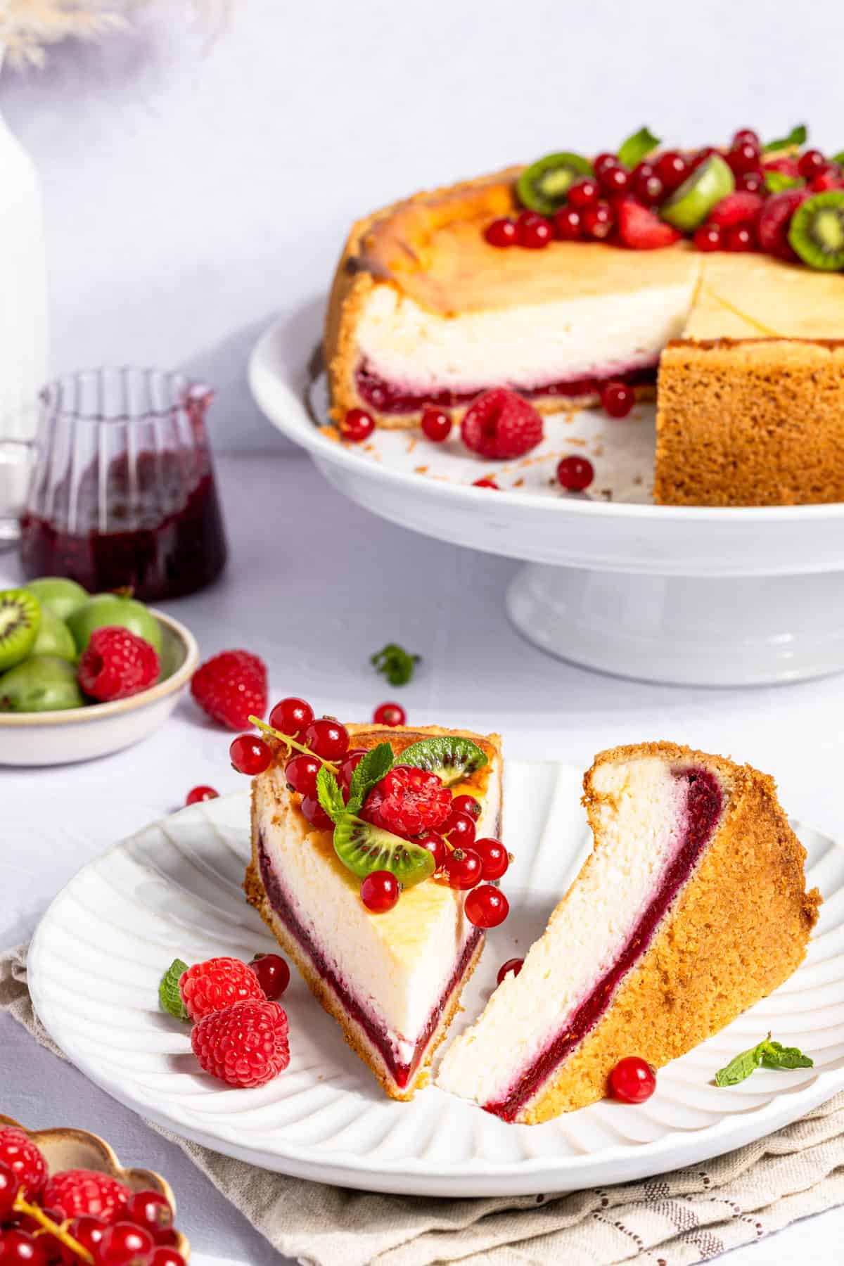 Ein Teller mit einem aufgeschnittenen Stück cremigem Käsekuchen mit Himbeerfüllung. Das Stück Kuchen zeigt deutlich die zweifarbige Schichtung: eine helle Quarkmasse und eine leuchtend rote Schicht aus Himbeerpüree am Boden. Der Kuchen mit Himbeerboden ist auf einem Teller mit frischen Himbeeren, roten Johannisbeeren und kleinen, grünen Mini-Kiwi-Beeren drapiert. Im Hintergrund steht der angeschnittene, ganze Cheesecake auf einer weißen Etagere.