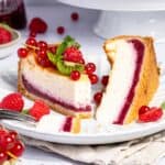 Ein Teller mit einem aufgeschnittenen Stück cremigem Käsekuchen mit Himbeerboden. Das Stück Kuchen zeigt deutlich die zweifarbige Schichtung: eine helle Quarkmasse und eine leuchtend rote Schicht aus Himbeerpüree am Boden. Der Kuchen mit Himbeerboden ist auf einem Teller mit frischen Himbeeren, roten Johannisbeeren und kleinen, grünen Mini-Kiwi-Beeren drapiert.