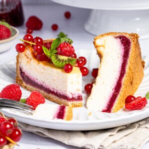 Ein Teller mit einem aufgeschnittenen Stück cremigem Käsekuchen mit Himbeerboden. Das Stück Kuchen zeigt deutlich die zweifarbige Schichtung: eine helle Quarkmasse und eine leuchtend rote Schicht aus Himbeerpüree am Boden. Der Kuchen mit Himbeerboden ist auf einem Teller mit frischen Himbeeren, roten Johannisbeeren und kleinen, grünen Mini-Kiwi-Beeren drapiert.