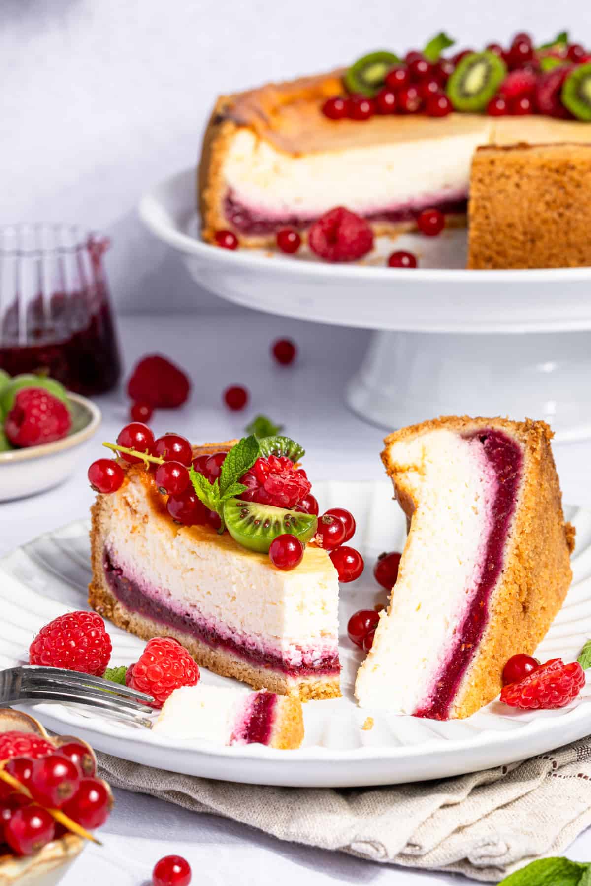 Ein Teller mit einem angeschnittenen Stück ultra cremigem Käsekuchen mit Himbeerboden, belegt mit frischen Himbeeren, roten Johannisbeeren und Mini-Kiwiberrys. Im Hintergrund ist der angeschnittene, ganze Käsekuchen auf einer weißen Etagere zu sehen, ebenfalls dekoriert mit Beeren und Minze. Der Himbeer-Cheesecake zeigt seine deutlich sichtbare, rote Fruchtschicht im Boden.