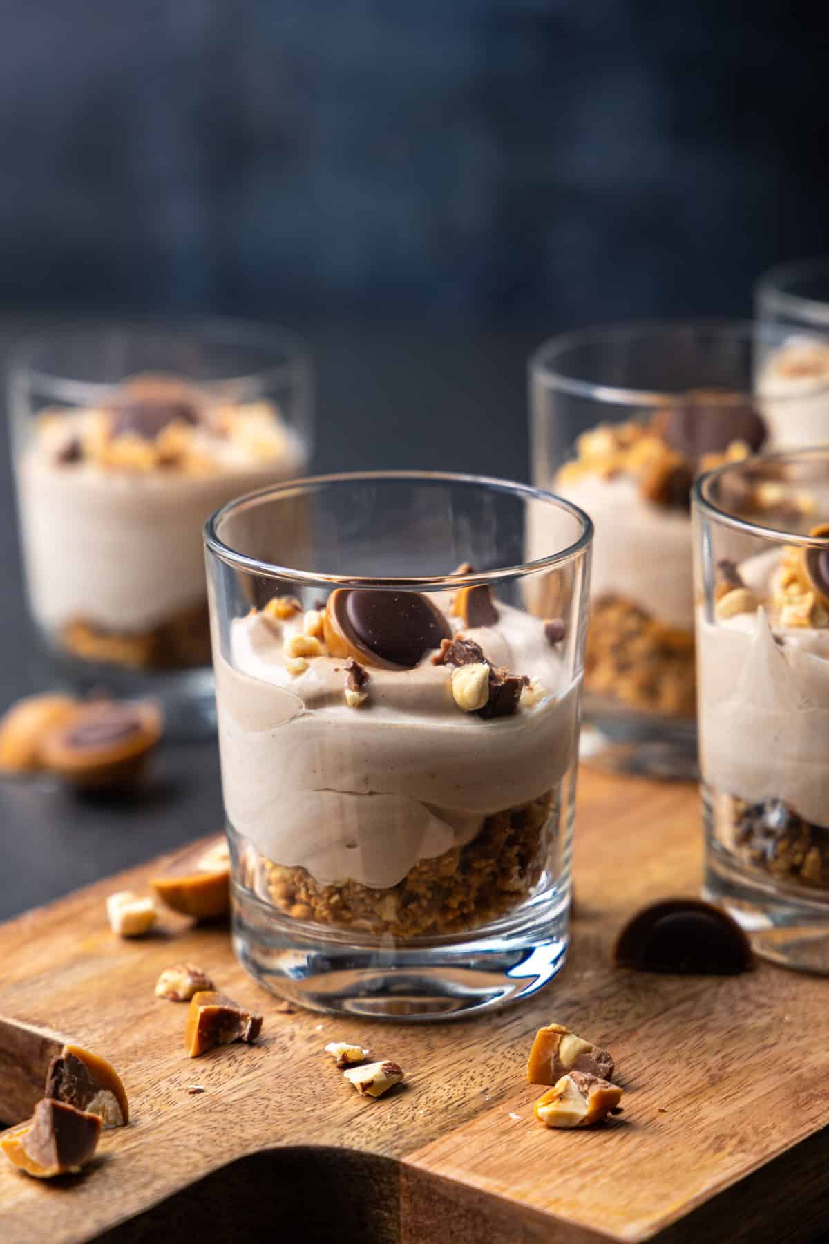 Nahaufnahme von sechs Toffifee-Desserts im Glas, arrangiert auf einem rustikalen Holzbrett und einem dunklen Hintergrund. Im Vordergrund steht ein Glas, das einen geschichteten Aufbau zeigt: unten ein goldbrauner, knuspriger Butterkeksboden, darüber eine hellbraune, cremige Toffifee-Creme. Das Dessert ist mit gehackten Haselnüssen und einem halben Toffifee garniert.
