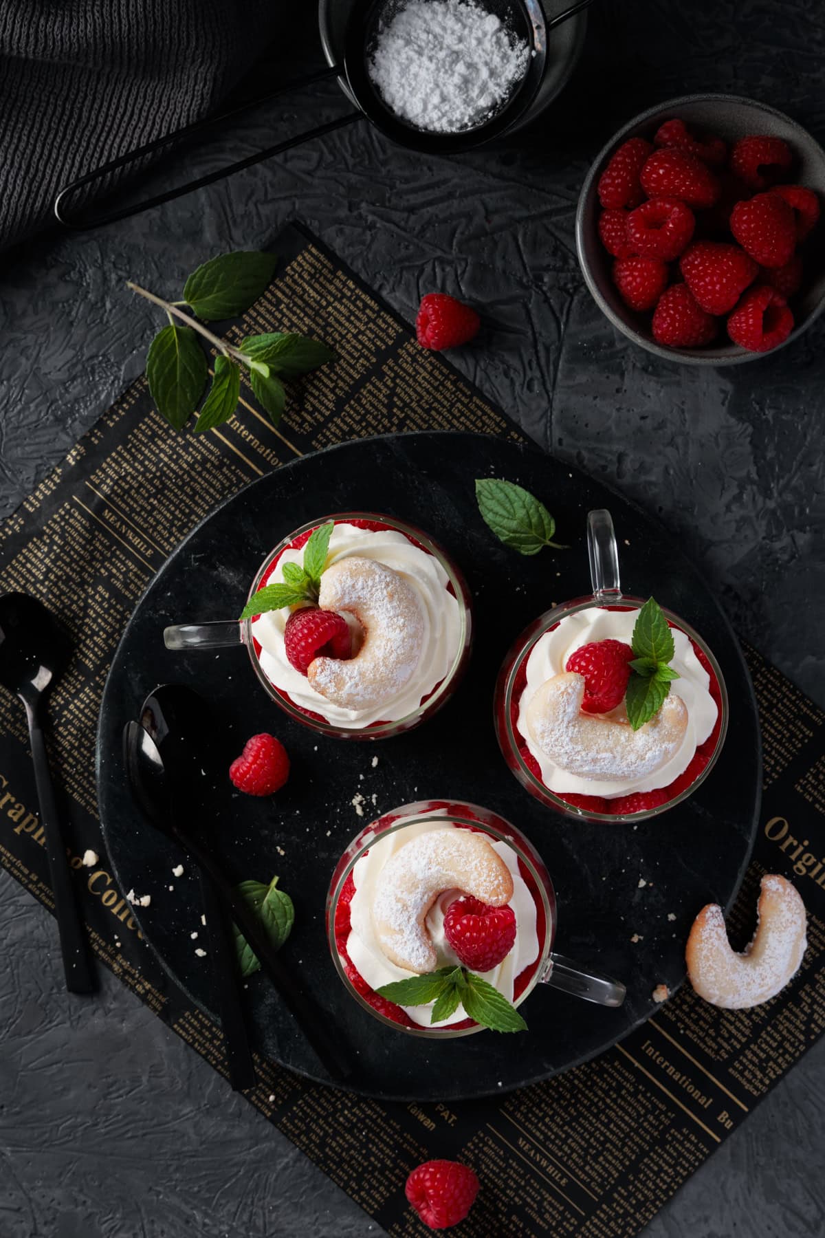 Drei Vanillekipferl-Desserts im Glas, von oben fotografiert. Die Desserts stehen auf einer dunklen Marmorplatte. Jedes Glas ist mit Himbeeren, einer weißen Creme und einer ganzen Vanillekipferl-Dekoration geschichtet. Einige frische Himbeeren und Minzblätter sind um die Gläser herum auf dem Tisch verteilt. Im Hintergrund sind eine Schale mit Himbeeren und eine Schüssel mit Puderzucker zu sehen. Das Bild zeigt ein festliches Schicht-Dessert für Weihnachten oder die Adventszeit aus der Vogelperspektive.