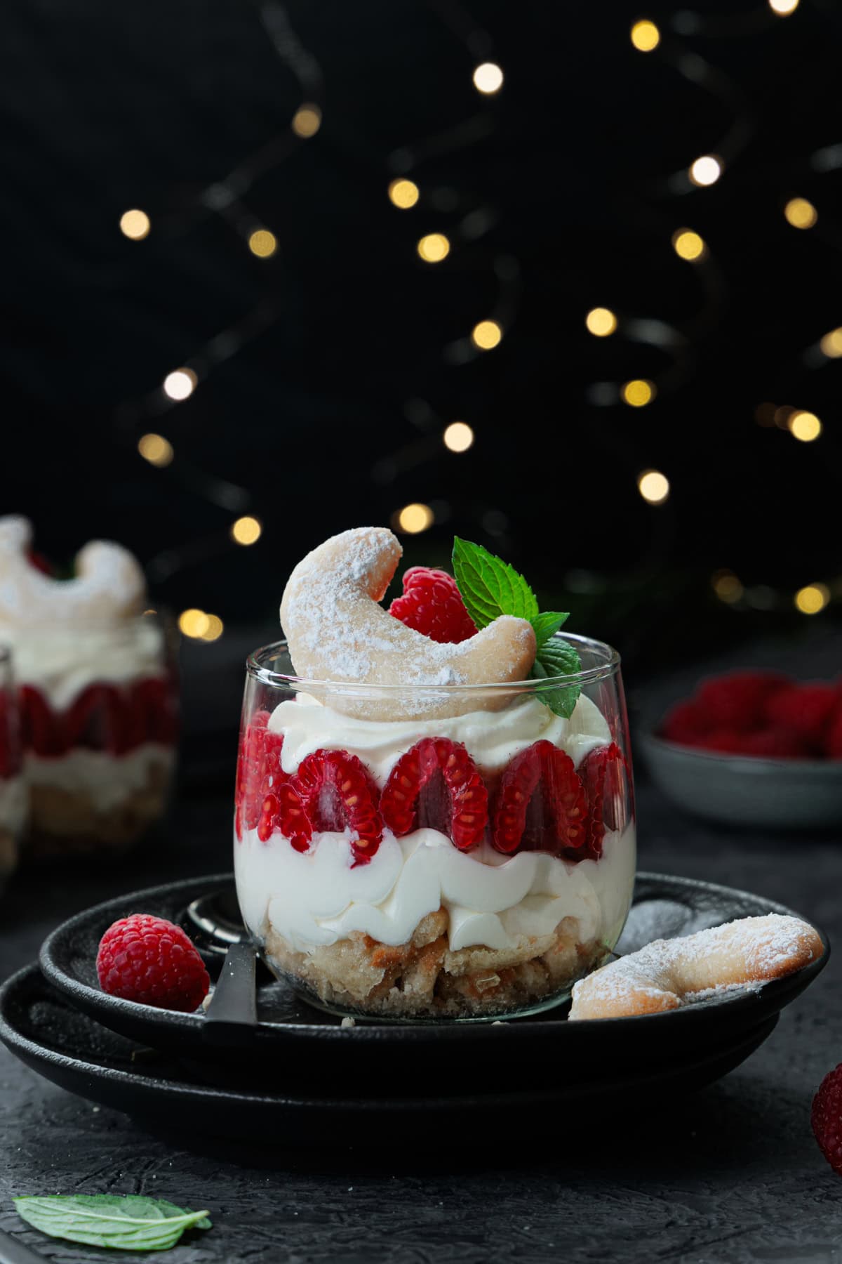 Ein appetitliches Vanillekipferl-Dessert im Glas steht auf einem dunklen Teller. Das Dessert ist in einem transparenten Glas geschichtet. Man sieht eine untere Schicht aus zerkleinerten Vanillekipferl, gefolgt von einer weißen, cremigen Schicht aus Mascarpone-Creme. Darauf sind rote, halbierte Himbeeren entlang des Glasrandes angeordnet. Die oberste Schicht ist eine weitere Portion der weißen Creme, die wie ein Swirl eingespritzt wurde. Obenauf liegt eine einzelne ganze Himbeere, ein halbes Vanillekipferl und ein frisches Minzblatt als Dekoration. Im Hintergrund sind weitere Gläser des Desserts unscharf zu sehen, ebenso wie eine kleine Schale mit frischen Himbeeren. Der gesamte Hintergrund ist dunkel und mit sanft leuchtenden, unscharfen Lichterketten dekoriert, was eine festliche, warme Atmosphäre schafft. 