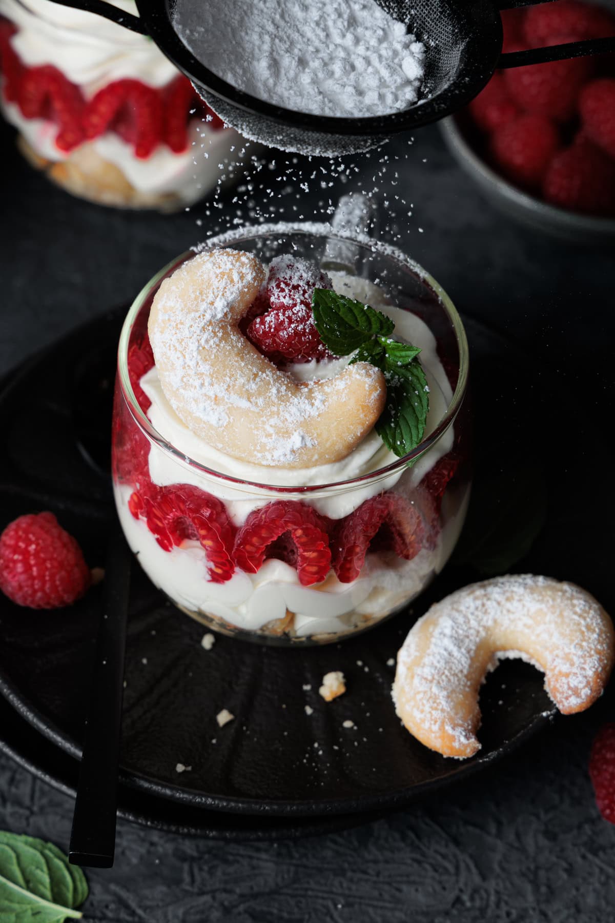Ein appetitlich angerichtetes Vanillekipferl-Dessert mit Himbeeren wird im Vordergrund mit Puderzucker bestäubt. Das Dessert befindet sich in einem runden Glas auf einem dunklen Teller. Man erkennt die sichtbaren Schichten: Eine Basis aus zerbröselten Kipferln, darüber eine helle, cremige Füllung, dann eine Schicht aus halbierten, leuchtend roten Himbeeren, die sich an der Glaswand anlehnen, gefolgt von einer weiteren großzügigen Schicht der Creme. Obenauf krönt ein Vanillekipferl, eine Himbeere und ein Minzblatt die Kreation. Eine Hand hält ein Sieb, aus dem feiner Puderzucker auf das Gericht herabrieselt und es wie mit frisch gefallenem Schnee überzieht. Im Hintergrund sind weitere Dessertgläser und eine Schale mit Himbeeren unscharf zu sehen, was die Szene sehr einladend wirken lässt.