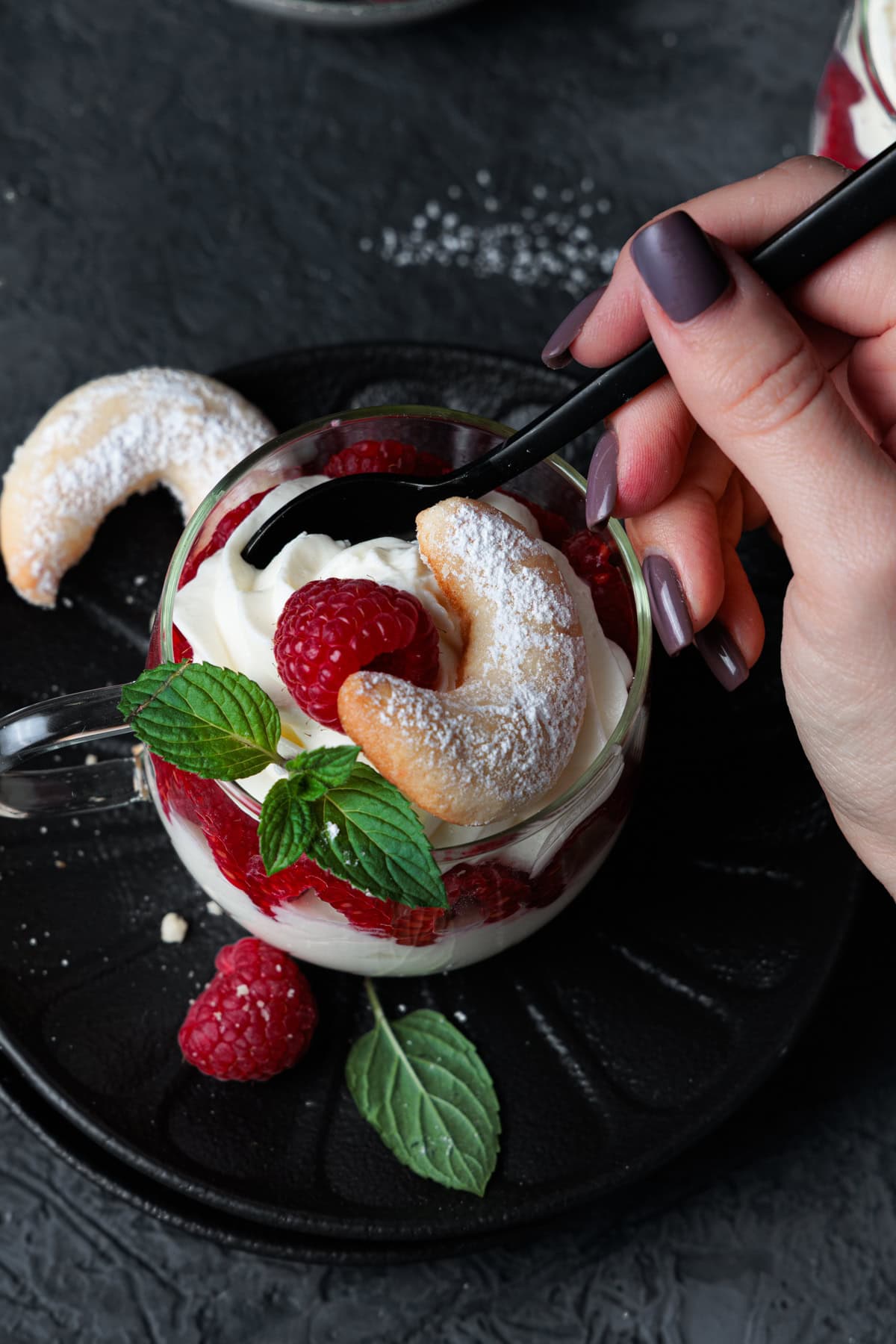Eine weibliche Hand hält einen schwarzen Löffel über ein einladendes Vanillekipferl-Dessert im Glas. Das festliche Dessert für Weihnachten ist kunstvoll geschichtet: Unten eine Schicht aus zerkleinerten Vanillekipferl, gefolgt von einer hellen, sahnigen Creme, darauf leuchtend rote Himbeeren und eine weitere Cremeschicht. Ein mit Puderzucker bestäubtes Vanillekipferl, eine frische Himbeere und ein Minzblatt schmücken die Oberfläche. Das Glas steht auf einem dunklen, geriffelten Teller. Rechts im Bildrand sind einzelne Himbeeren und Minzblätter zu sehen, die auf einer dunkelgrauen, leicht strukturierten Oberfläche liegen. Die Gesamtkomposition lädt dazu ein, dieses einfache Weihnachtsdessert sofort zu genießen.