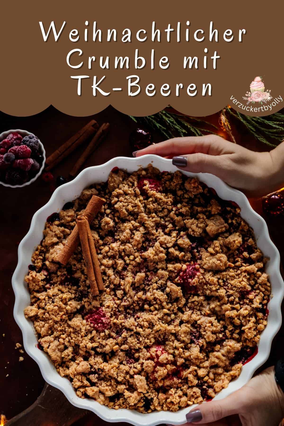 Pinterest-Pin: Ein von zwei Händen gehaltener, frisch gebackener weihnachtlicher Crumble mit TK-Beeren in einer runden, weißen Backform, bereit zum Servieren. Die knusprigen Streusel sind goldbraun und mit Zimtstangen dekoriert. Der Hintergrund zeigt verschwommene weihnachtliche Dekorationen wie Tannenzweige, Beeren und Lichterketten. Das Bild trägt den Schriftzug "Weihnachtlicher Crumble mit TK-Beeren" und das Logo "verzuckertbyolly".