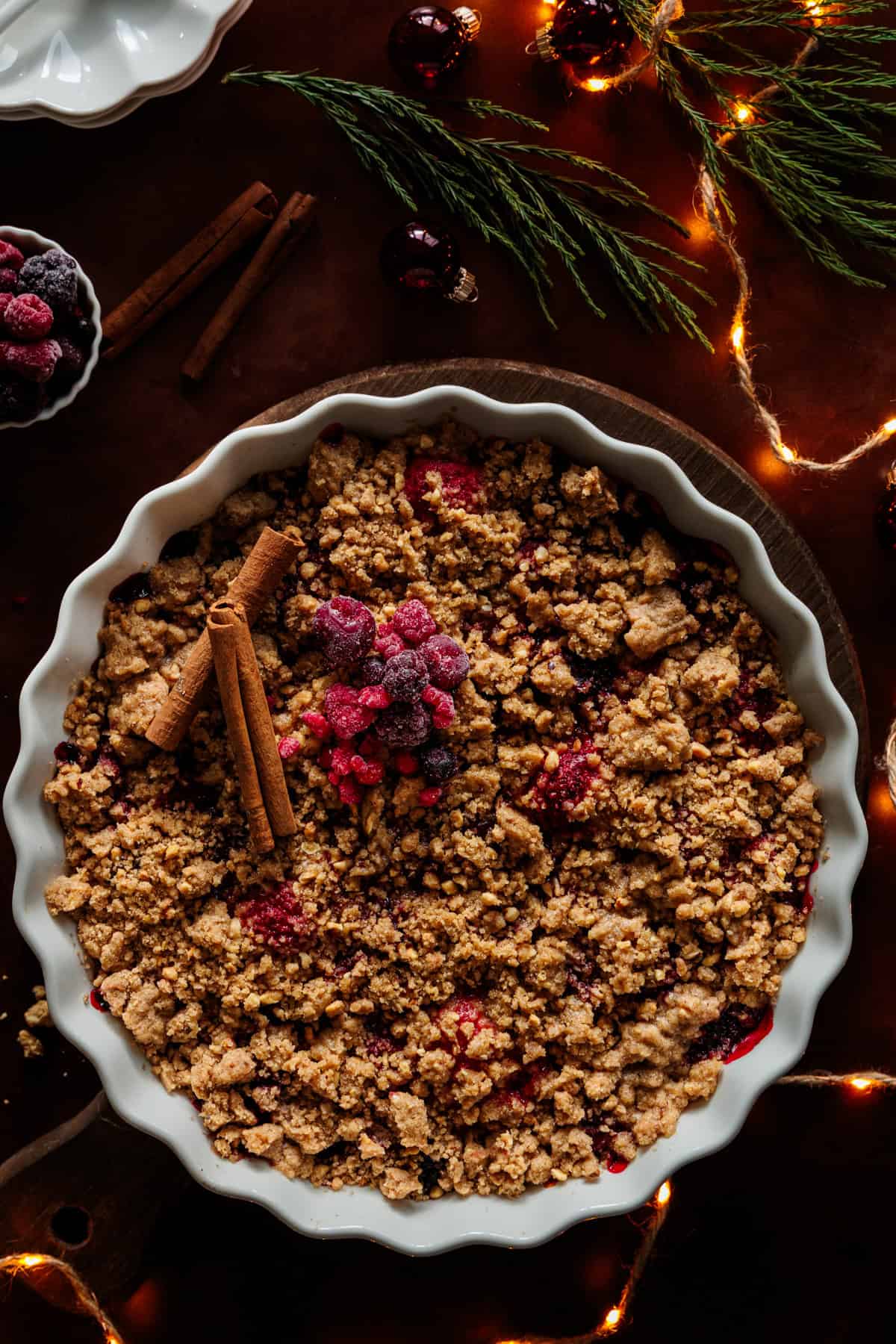 Ein festlicher TK-Beeren-Crumble in einer weißen, runden Auflaufform, bestreut mit knusprigen Streuseln und dekoriert mit Zimtstangen und einigen TK-Beeren. Im Hintergrund sind weihnachtliche Dekorationen wie Tannenzweige, Lichterketten und rote Christbaumkugeln zu sehen, die eine warme und gemütliche Atmosphäre schaffen.