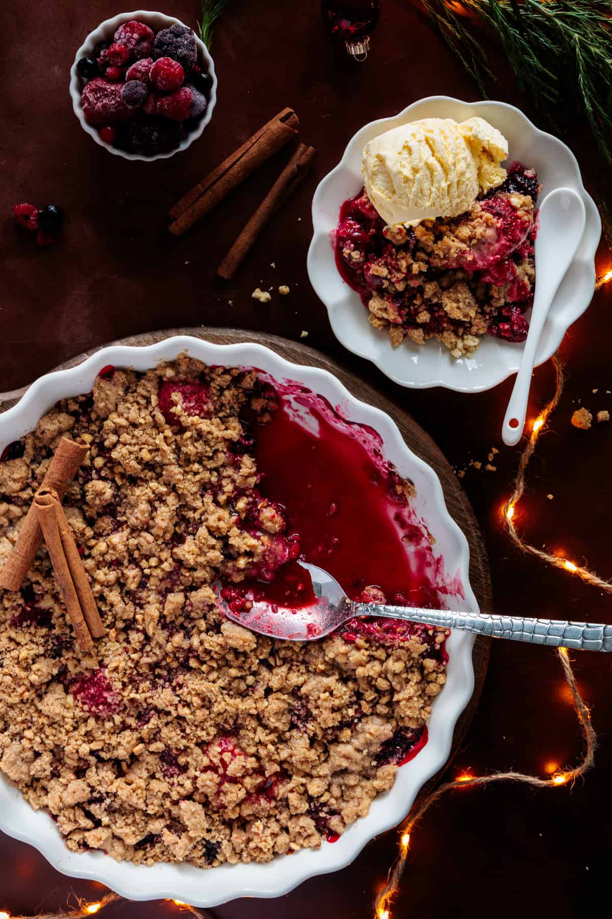 Nahaufnahme eines Beeren-Crumbles in einer weißen, runden Auflaufform. Eine kleine Portion des Weihnachts-Crumbles liegt mit einer Kugel Vanilleeis auf einem separaten, kleineren Teller daneben. Zimtstangen, eine Schüssel mit gefrorenen Beeren und Lichterketten sind ebenfalls im Bild, was eine gemütliche und festliche Atmosphäre erzeugt.