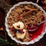 Ein weihnachtlicher Beeren-Crumble in einer weißen Form, der gerade serviert wird. Ein Löffel liegt in der warmen, saftigen Fruchtfüllung und zwei Kugeln Vanilleeis, garniert mit frischen TK-Beeren, liegen auf den knusprigen Streuseln. Das Bild zeigt weihnachtliche Dekorationen wie Zimtstangen, Lichterketten und Tannenzweige.
