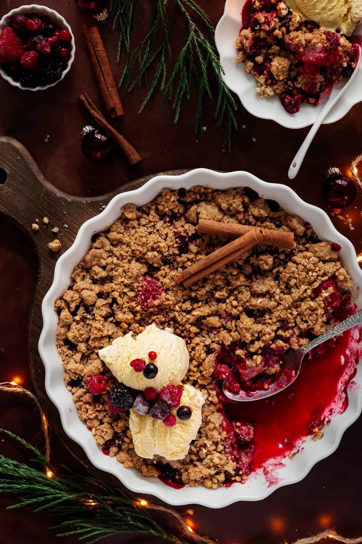  Ein weihnachtlicher Beeren-Crumble in einer weißen Form, der gerade serviert wird. Ein Löffel liegt in der warmen, saftigen Fruchtfüllung und zwei Kugeln Vanilleeis, garniert mit frischen TK-Beeren, liegen auf den knusprigen Streuseln. Das Bild zeigt weihnachtliche Dekorationen wie Zimtstangen, Lichterketten und Tannenzweige.