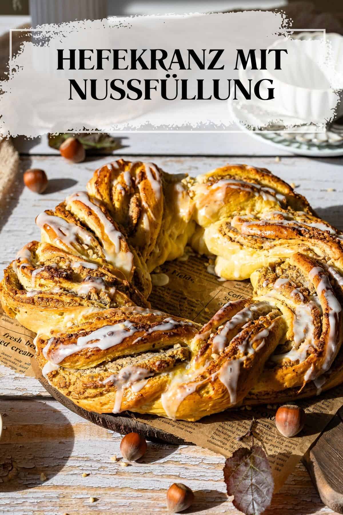 Pin zu Pinterest: Ganze Aufnahme des fertigen, geflochtenen Nusskranzes (Hefekranz mit Nussfüllung aus dem Thermomix®), der mit weißer Glasur besprenkelt ist und auf einer rustikalen Holzoberfläche liegt. Der Kranz ist von einzelnen Haselnüssen und getrockneten Blättern umgeben. Starkes, seitliches Sonnenlicht hebt die goldbraune Oberfläche und die Textur des Gebäcks hervor.

