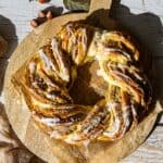 Ganze Aufnahme des fertigen, geflochtenen Nusskranzes (Hefekranz mit Nussfüllung aus dem Thermomix®), der mit weißer Glasur besprenkelt ist und auf einer rustikalen Holzoberfläche liegt.