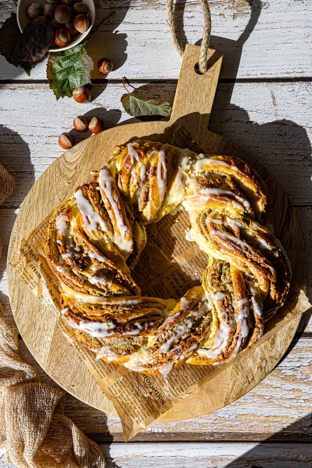 Herbstlicher Nusskranz / Hefekranz mit Nussfüllung aus dem Thermomix®. Der geflochtene Kranz mit heller Zuckerglasur liegt auf einem Holzbrett. Im Hintergrund sind herbstliche Dekoration, Haselnüsse und Blätter zu sehen. Das Rezept ist perfekt für Backen im Herbst und Winter.