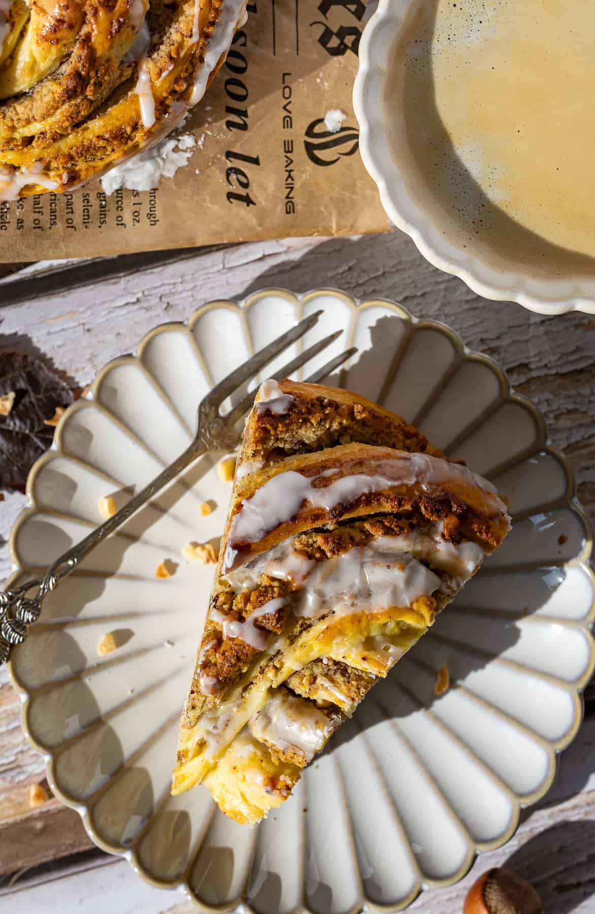 Ein dreieckiges Stück vom Nusskranz / Hefekranz mit dicker Nussfüllung und weißer Zuckerglasur wird auf einem kleinen, weißen Kuchenteller mit goldener Verzierung und einer Gabel präsentiert. Oben rechts ist eine Tasse Kaffee oder Milchkaffee zu sehen.