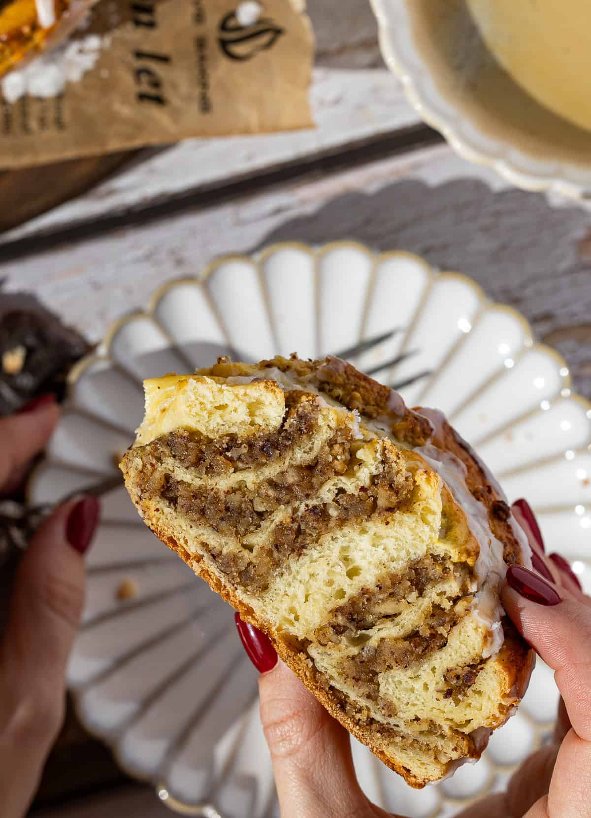 Nahaufnahme eines Anschnitts vom gebackenen Nusskranz Hefekranz. Man sieht deutlich die saftige Schichtung von hellem Hefeteig und dunkler Nussfüllung, überzogen mit weißer Zuckerglasur. Das Teigstück wird von Händen mit dunklem Nagellack gehalten und liegt auf einem verzierten Kuchenteller.