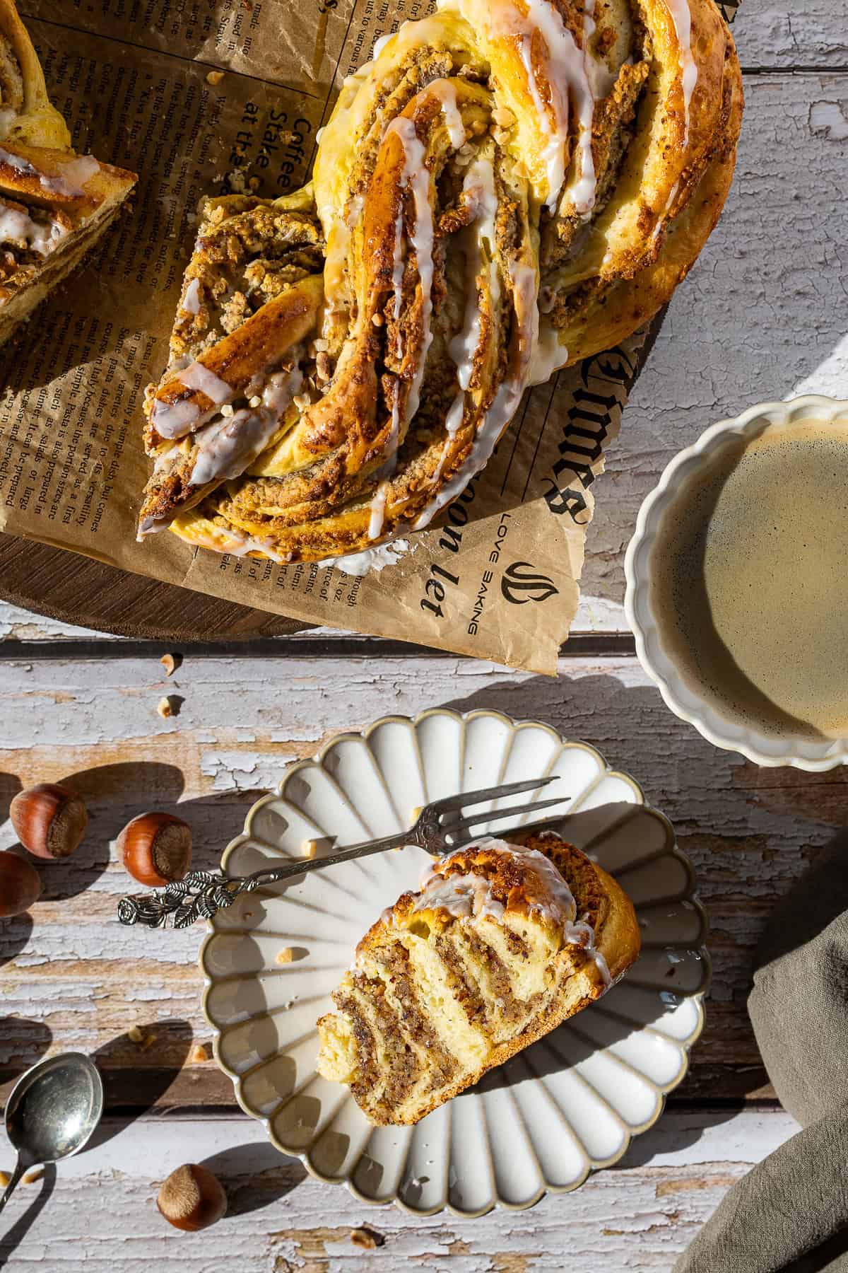 Ansicht von oben (Top View) des Nusskranzes aus dem Thermomix®. Ein einzelnes, saftiges Stück liegt auf einem kleinen, verzierten Kuchenteller mit einer Gabel. Im Hintergrund ist der restliche, angeschnittene Kranz auf braunem Papier zu sehen, zusammen mit einer Tasse Kaffee, ganzen Haselnüssen und einem Löffel auf dem rustikalen Holztisch.
