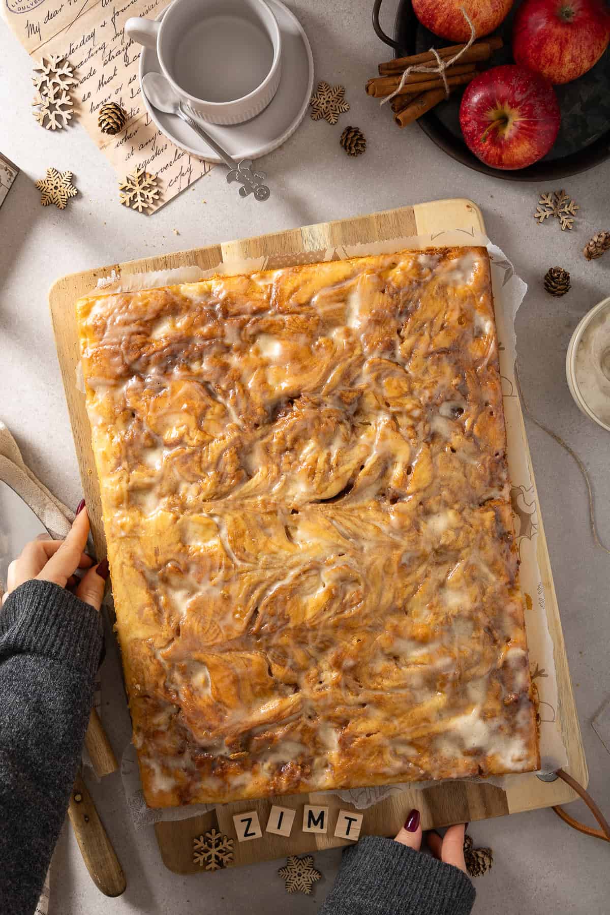 Frisch gebackener Apfel-Zimtschneckenkuchen vom Blech auf einem Holzbrett. Der saftige Kuchen zeigt eine goldbraune Zimt-Marmorierung mit Äpfeln und ist mit einem cremigen, weißen Crème-fraîche-Guss überzogen. Im Hintergrund sind herbstliche Details wie Zimtstangen, Äpfel und eine Tasse Kaffee zu sehen. Ein leckeres Thermomix® Rezept für Zimtschneckenkuchen.