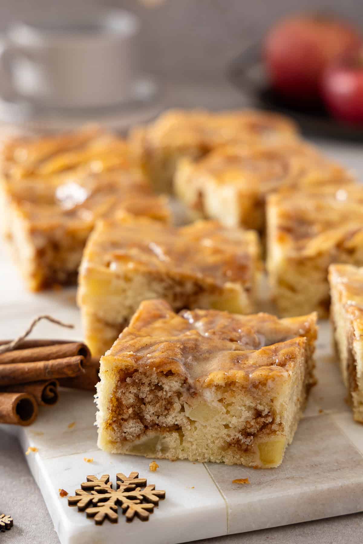 Frisch geschnittene Stücke Apfel-Zimtschneckenkuchen vom Blech, appetitlich angerichtet auf einer weißen Marmorplatte. Die goldbraune Zimt-Marmorierung und die saftigen Apfelstücke sind im Inneren der Kuchenstücke gut sichtbar, überzogen mit einer glänzenden Crème-fraîche-Glasur. Dekorativ daneben liegen Zimtstangen und eine hölzerne Schneeflocke. Ein ideales Herbstrezept und Weihnachtskuchen, einfach mit dem Thermomix® zubereitet.