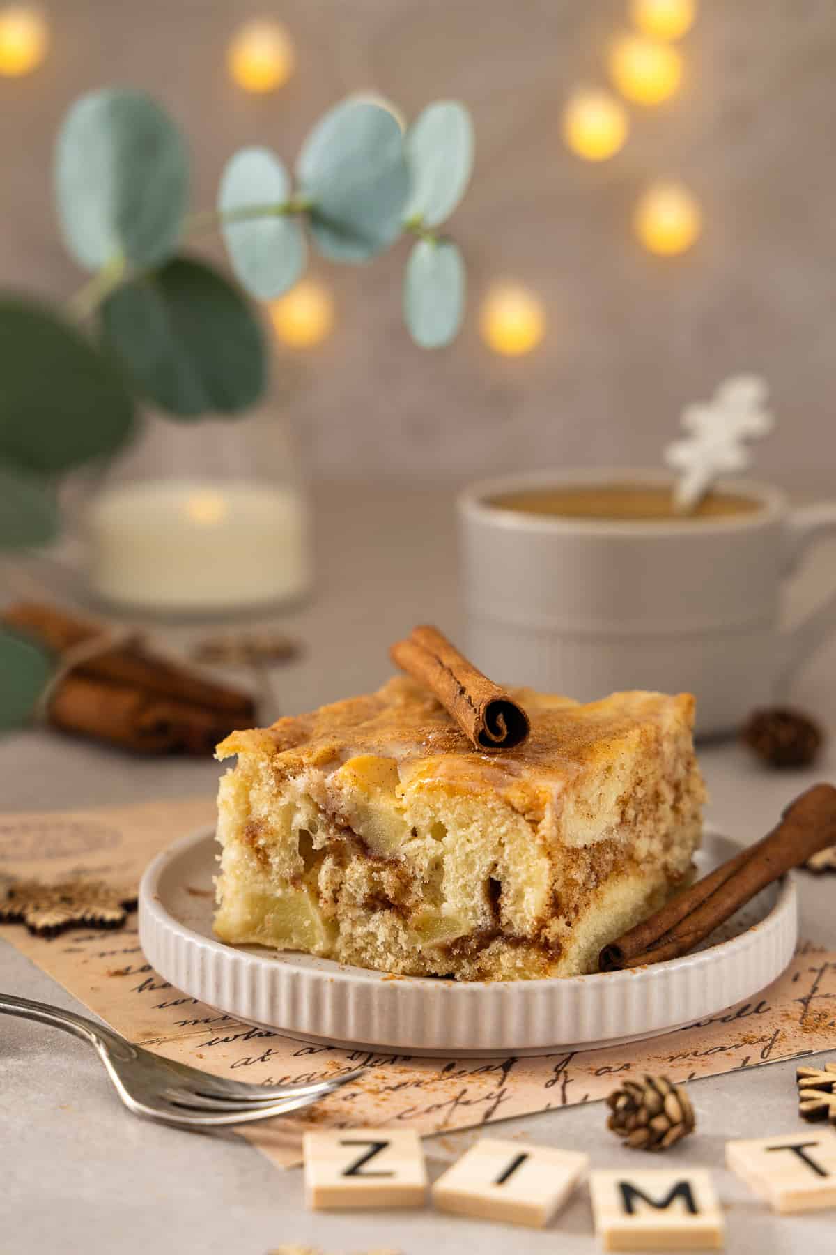 Nahaufnahme eines Stücks Apfel-Zimtschneckenkuchen vom Blech auf einem weißen Teller. Das saftige Kuchenstück zeigt deutlich die goldbraune Zimt-Marmorierung und Apfelstücke im Teig, verziert mit einer Zimtstange und einem Hauch Crème-fraîche-Guss. Im unscharfen Hintergrund sind herbstliche Dekorationen, Zimtstangen und eine Kaffeetasse zu sehen. Ein leckeres Thermomix® Rezept für Zimtschneckenkuchen.