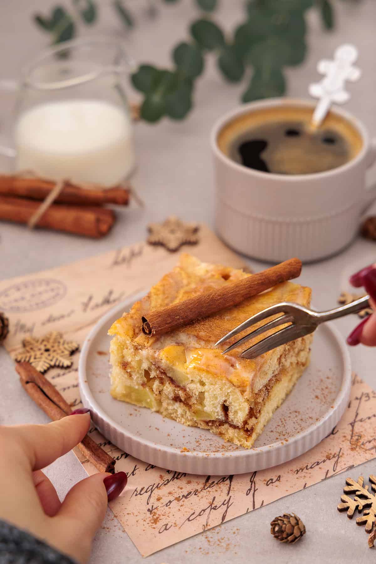 Eine Hand schneidet mit einer Gabel ein Stück saftigen Apfel-Zimtschneckenkuchen vom Blech an, der auf einem hellen Teller liegt. Die goldbraune Zimt-Marmorierung und die weichen Apfelstücke im Kuchen sind deutlich zu erkennen, überzogen mit einer Crème-fraîche-Glasur und einer Zimtstange als Dekoration. Im Hintergrund eine Tasse Kaffee, Milch und herbstliche Details.  Ein ideales Rezept für die Apfelsaison oder als Dessert für Weihnachten, ganz schnell und einfach mit dem Thermomix® zubereitet.
