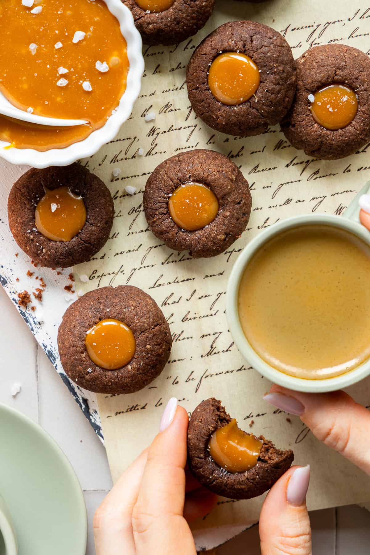Detaillierte Nahaufnahme von dunklen Schoko-Engelsaugen mit glänzender Karamell-Füllung und Salzkristallen (Salted Caramel) auf einem hellen Untergrund mit alter Schrift. Eine Hand hält einen angebissenen Keks, der den cremigen Karamell-Kern zeigt. Im Hintergrund stehen eine Tasse Espresso und eine Schale mit zusätzlicher Karamell-Füllung.
