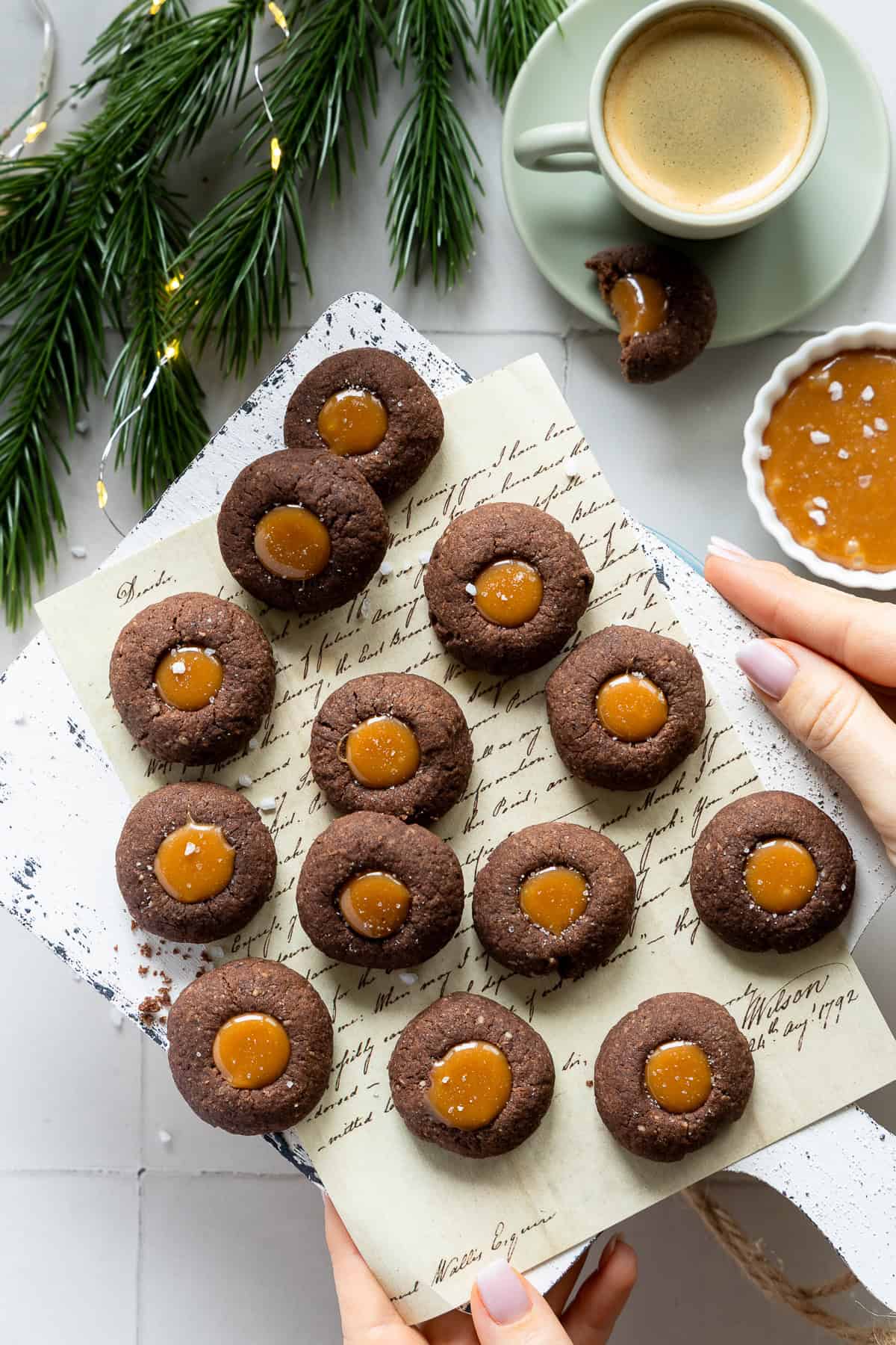 Etwa ein Dutzend runde Schoko-Engelsaugen, die auf einem rustikalen, weißen Holzbrett liegen, das mit einer handschriftlichen Notiz verziert ist. Die Plätzchen bestehen aus dunklem Schokomürbeteig und sind mit einer glänzenden, dicken Karamell-Füllung befüllt, die teilweise mit grobem Meersalz (Salted Caramel) bestreut ist. Im Hintergrund sind weihnachtliche Akzente wie grüne Tannenzweige, Lichterketten und zwei Kaffeetassen mit Espresso zu sehen. Eine Hand hält das Brett leicht fest, während der Fokus auf den saftigen Schoko-Keksen liegt, deren Füllung cremig aus der Mulde quillt.