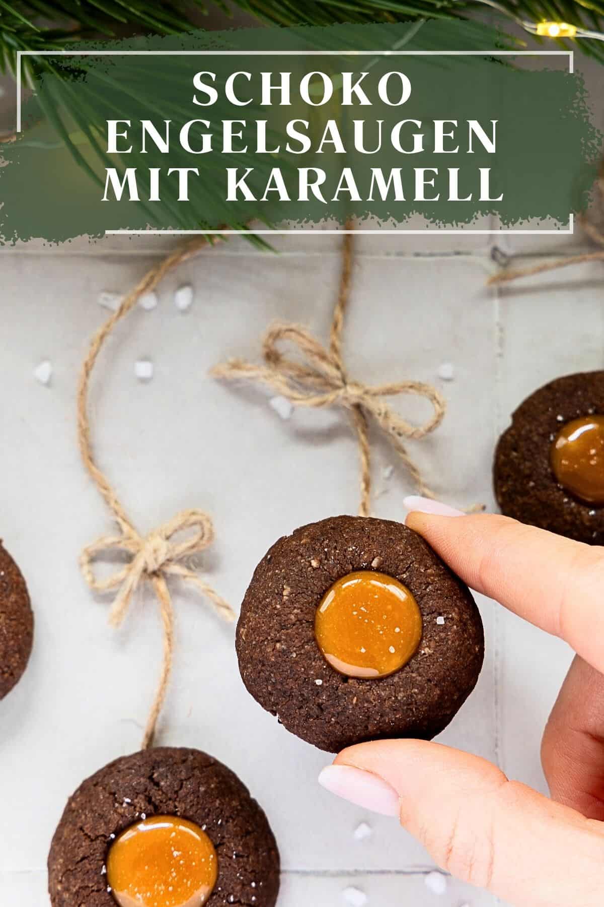 Ein Pinterest-Pin mit dem Titel "SCHOKO ENGELSAUGEN MIT KARAMELL" auf einem dunklen Banner oben. Das Bild zeigt eine Nahaufnahme einer Hand, die eine runde, dunkle Schoko-Engelsauge mit glänzender Karamell-Füllung und feinen Salzkristallen (Salted Caramel) hochhält. Im Hintergrund sind weitere, an Schnüren befestigte Plätzchen zu sehen, umgeben von Tannengrün und verstreuten Salzkristallen, was eine festliche, weihnachtliche Stimmung erzeugt.
