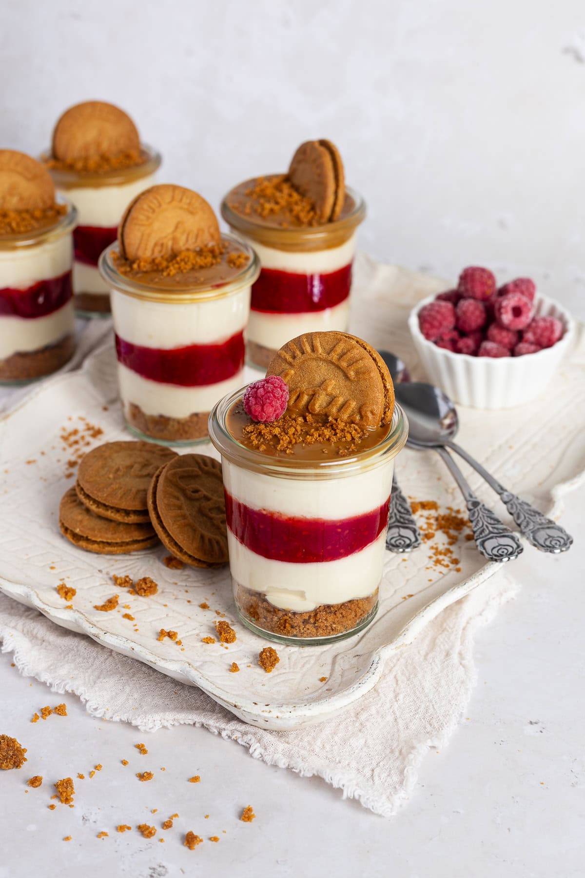 Fünf gefüllte Lotus Biscoff Himbeeren Desserts im Glas stehen auf einer hellen, strukturierten Servierplatte. Zu sehen sind die klaren Schichten: ein Keksboden, eine leuchtend rote Himbeerschicht und eine cremige No-Bake Quark-Joghurt-Creme. Die Gläser sind mit Lotus Keksen dekoriert. Daneben liegen weitere Kekse und lockere Krümel auf der Platte.