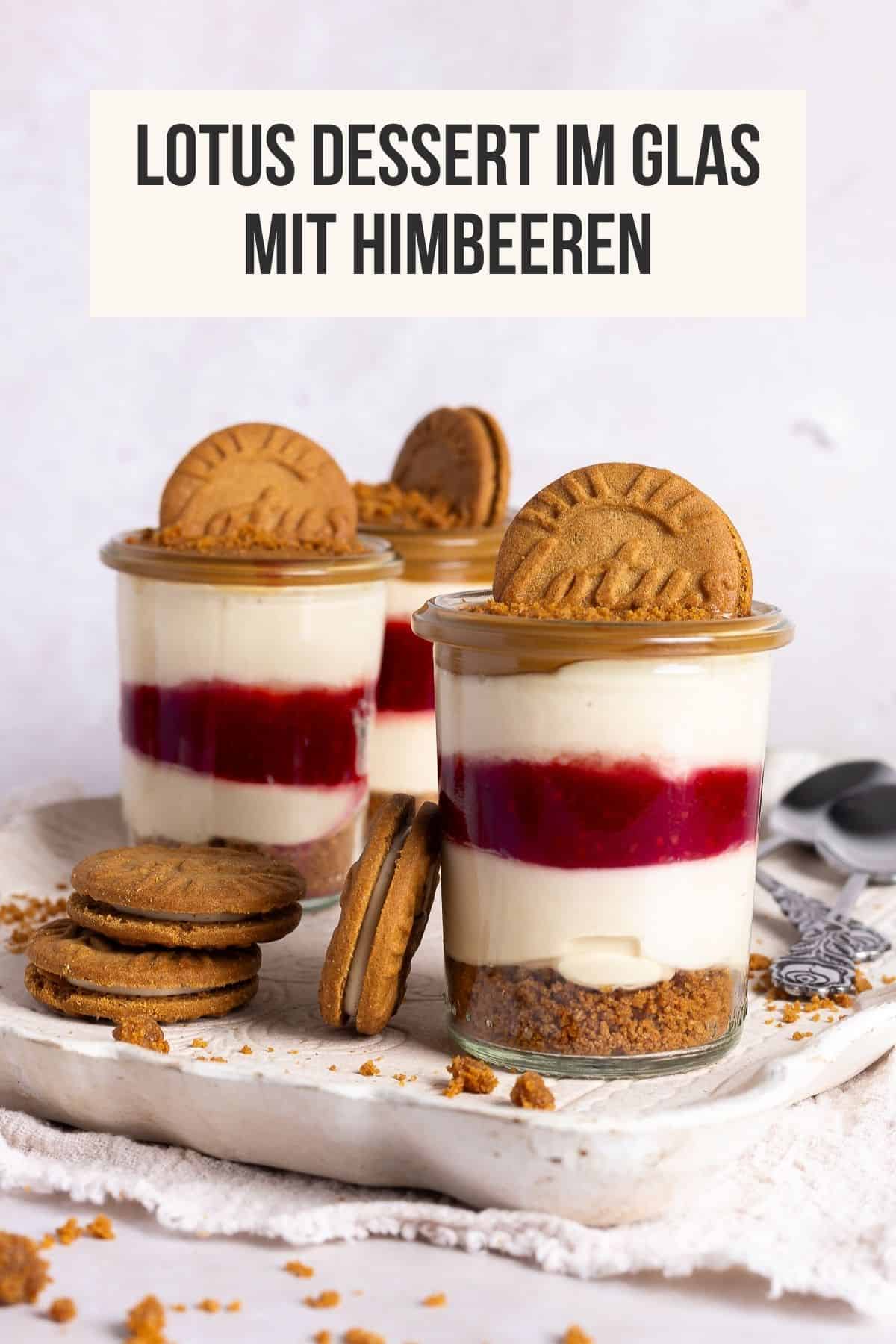 Pin für Pinterest: Drei Lotus Biscoff Himbeeren Desserts im Glas stehen auf einem weißen Tablett unter einem Banner mit dem Titel "Lotus Dessert im Glas mit Himbeeren". Das cremige No-Bake Schichtdessert zeigt die klare Schichtung des Keksbodens, der roten Himbeerschicht und der Quark-Joghurt-Creme, dekoriert mit Lotus Keksen und Krümeln.