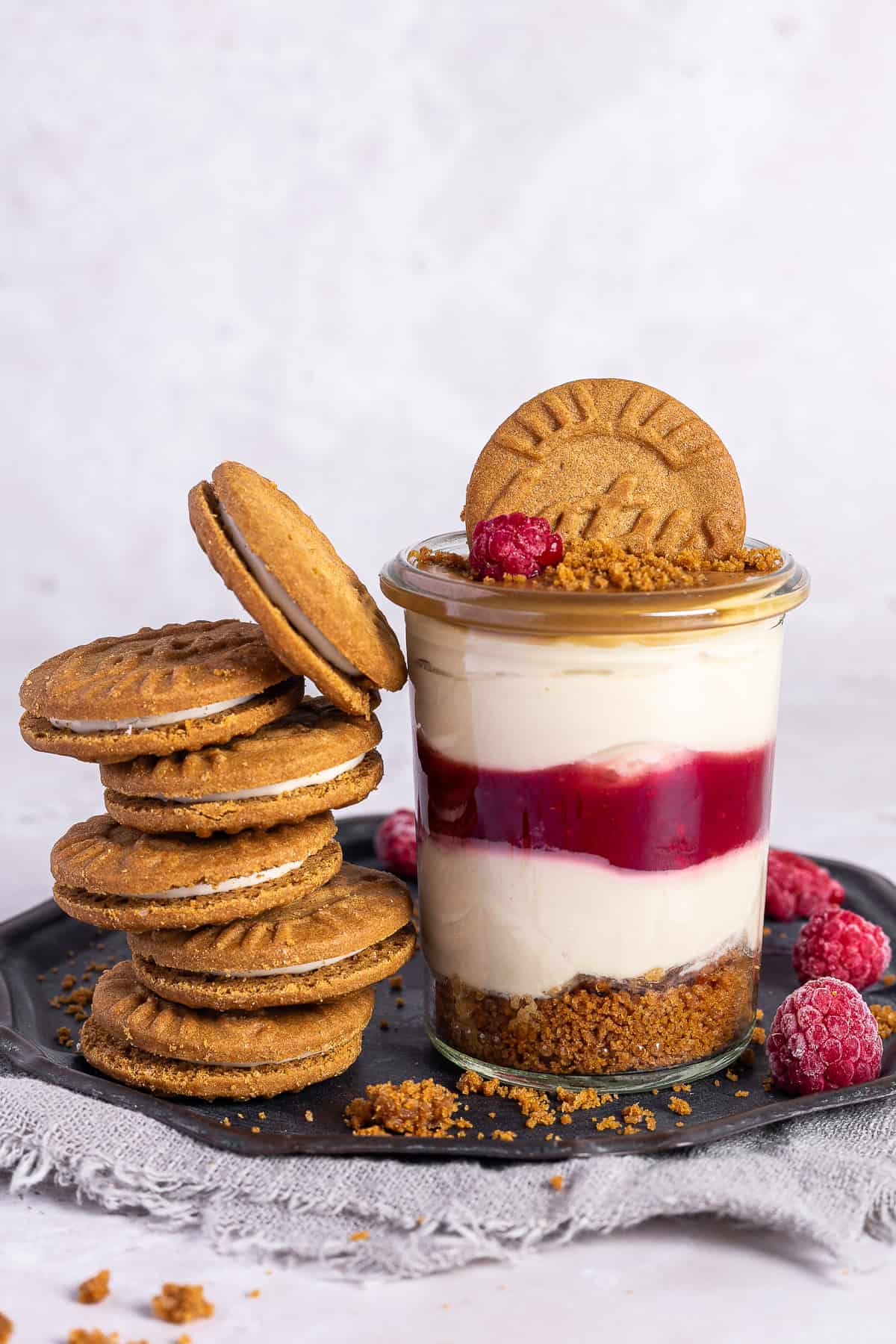 Ein einzelnes Lotus Biscoff Himbeeren Dessert im Glas steht auf einer dunklen Platte. Das cremige No-Bake Schichtdessert zeigt den Biscoff Keksboden, eine rote Himbeerschicht und die weiße Quark-Joghurt-Creme. Links neben dem Glas ist ein hoher Stapel Lotus Biscoff Sandwich-Kekse gestapelt. Das Glas ist mit einem ganzen Keks und frischen Himbeeren dekoriert.