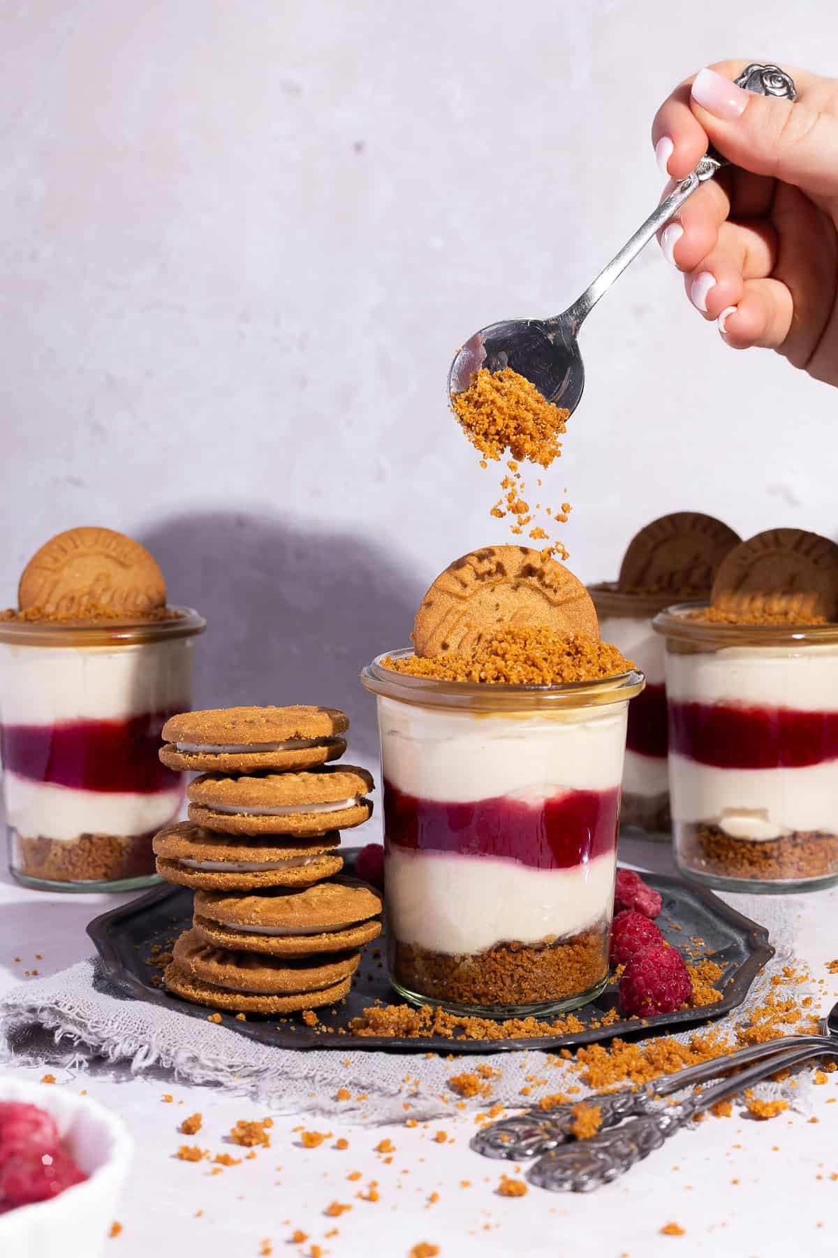 Die Hand einer Person bestreut ein Lotus Biscoff Himbeeren Dessert im Glas mit zerbröselten Lotus Biscoff Keksen von einem Löffel. Im Vordergrund steht das cremige No-Bake Schichtdessert mit Keksboden, Himbeerschicht und Creme auf einer dunklen Platte. Im Hintergrund sind drei weitere Gläser und ein Stapel Lotus Sandwich-Kekse zu sehen.