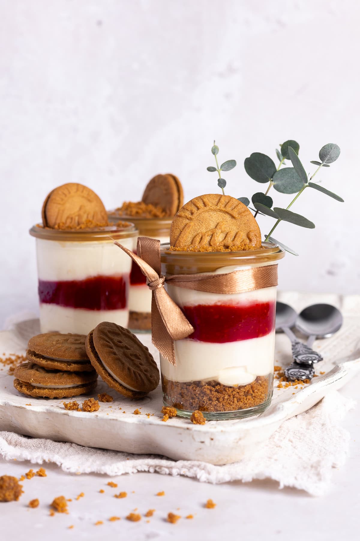 Drei festlich dekorierte Lotus Biscoff Himbeeren Desserts im Glas stehen auf einer hellen Platte. Das cremige No-Bake Schichtdessert zeigt die Schichten von Keksboden, roter Himbeerschicht und Quark-Joghurt-Creme. Das vordere Glas ist mit einem braunen Satinband und einem kleinen Eukalyptuszweig dekoriert, ideal für das Weihnachtsmenü.