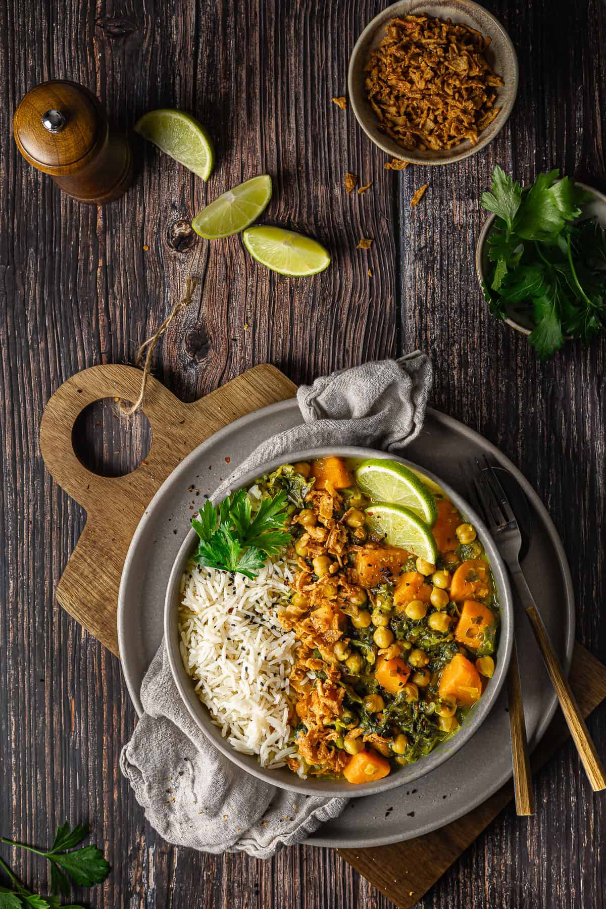 Flatlay-Aufnahme einer Schale mit veganem Süßkartoffel-Spinat-Curry, Kichererbsen und Basmati-Reis mit Schwarzkümmel, angerichtet auf einem dunklen Holztisch. Die Schale ist mit Röstzwiebeln, Petersilie und Limette garniert. Daneben liegen eine separate Schale mit Röstzwiebeln, frische Petersilie, Limettenviertel, eine Pfeffermühle und Besteck. Ein einfaches und schnelles Thermomix® Rezept für den Veganuary.