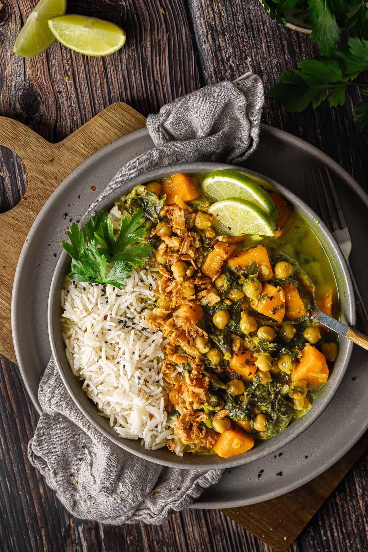 Zwei Hände in einem braunen Strickpullover halten eine Schale mit veganem Süßkartoffel-Spinat-Curry und Kichererbsen. Das cremige Gericht ist mit knusprigen Röstzwiebeln, frischer Petersilie und Limettenscheiben garniert und wird neben Basmati-Reis mit Schwarzkümmel serviert. Ein schnelles, wärmendes Thermomix® Rezept, ideal für den Veganuary.