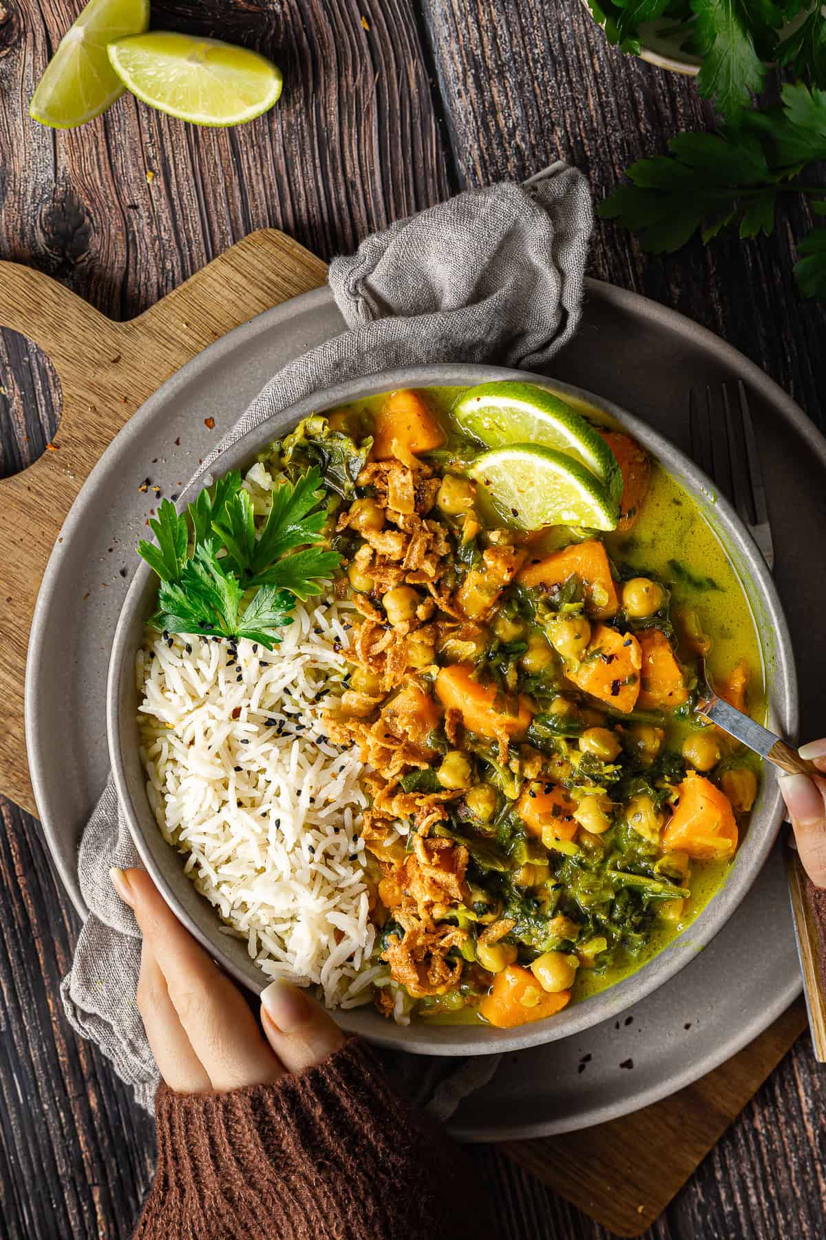 Draufsicht auf eine Schale mit cremigem veganen Süßkartoffel-Spinat-Curry und Kichererbsen, angerichtet neben Basmati-Reis mit Schwarzkümmel. Das Gericht ist garniert mit knusprigen Röstzwiebeln, frischer Petersilie und Limettenscheiben. Zwei Hände in einem braunen Strickpullover halten die warme Schüssel. Ein schnelles und einfaches Thermomix® Rezept, perfekt für den Veganuary.