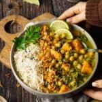 Flatlay-Ansicht von veganem Süßkartoffel-Spinat-Curry mit Kichererbsen und Basmati-Reis mit Schwarzkümmel. Die Schale steht auf einem dunklen Holztisch und ist mit knusprigen Röstzwiebeln, frischer Petersilie und Limettenscheiben garniert. Umgeben von Limettenspalten, einer Pfeffermühle und goldenem Besteck. Ein einfaches, schnelles Thermomix® Rezept für den Veganuary.