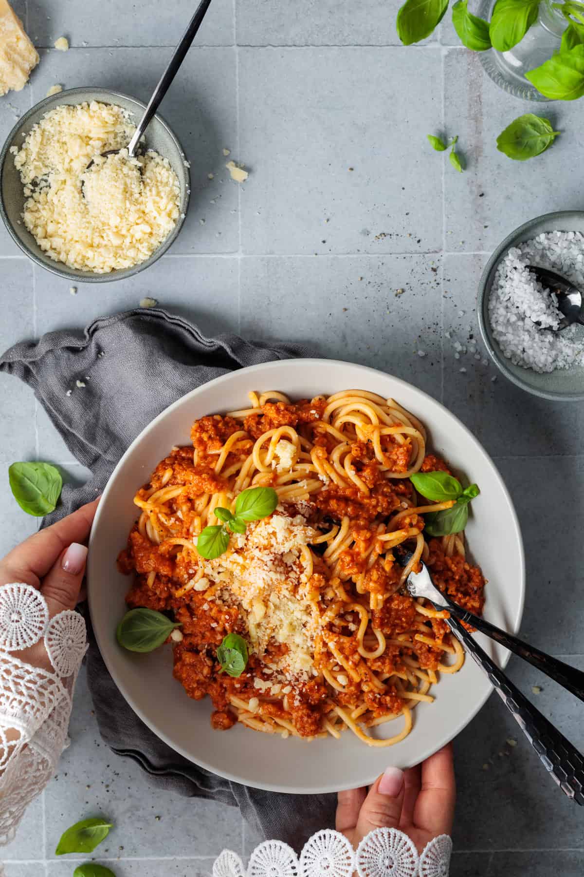 Draufsicht auf einen Teller Spaghetti mit vegetarischer Blumenkohl-Champignon-Bolognese aus dem Thermomix®, der von Händen in Spitzenärmeln gehalten wird. Das gesunde Pastagericht ist appetitlich mit Parmesan und Basilikum garniert und auf einem modernen grauen Untergrund neben Schälchen mit Meersalz und Käse angerichtet.