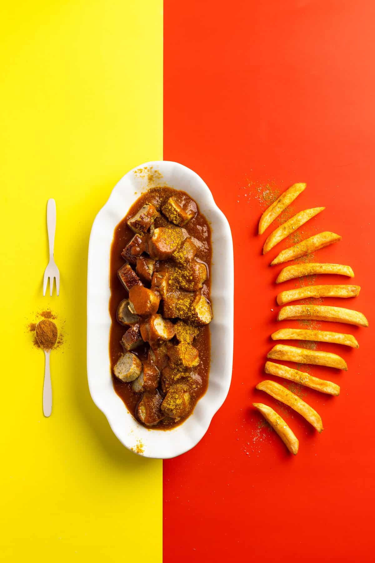 Eine Draufsicht auf eine klassische Currywurst in hausgemachter Soße, serviert in einer weißen Porzellanschale. Die Wurst ist mit Currypulver bestreut und liegt auf einem zweigeteilten Hintergrund in Gelb und Rot. Daneben sind knusprige Pommes frites kunstvoll in einer Kurve aufgereiht, ergänzt durch eine kleine Gabel und einen Löffel mit Currygewürz.