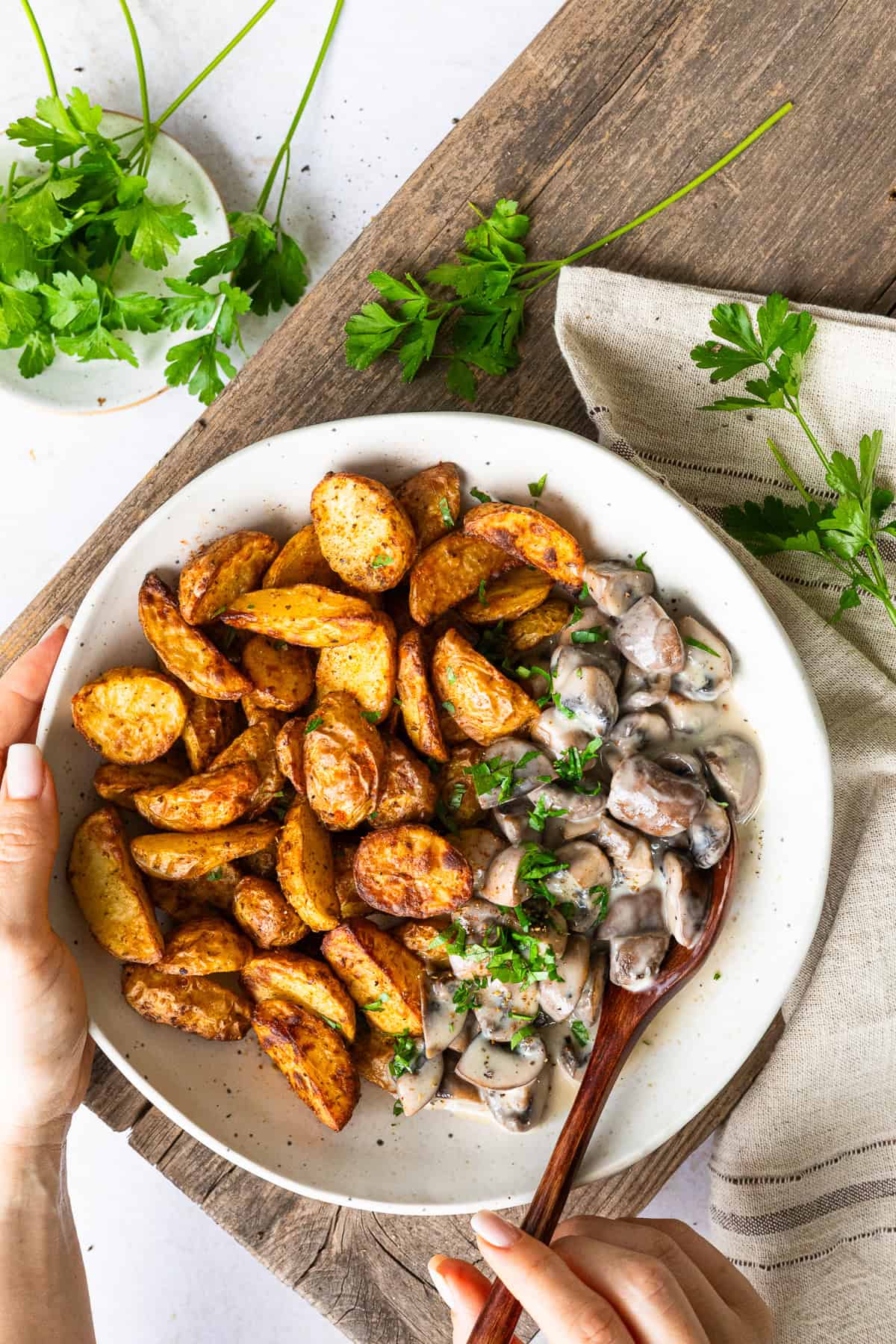 Zwei Hände halten eine weiße Schüssel mit einer Kartoffel-Champignon-Bowl auf einem rustikalen Holzbrett. Links liegen knusprig goldbraune Drillinge, rechts cremige Champignon-Rahmsoße mit frischer Petersilie. Ein Holzlöffel steckt in der Soße.