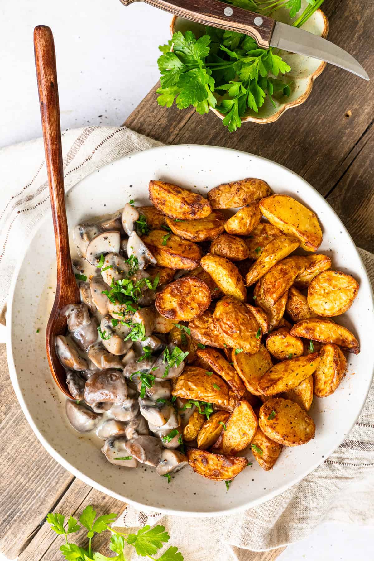 Eine helle Keramikschale mit knusprig goldbraunen Airfryer-Kartoffeln und einer cremigen Champignon-Rahmsoße, garniert mit frischer Petersilie auf einem rustikalen Holzhintergrund.