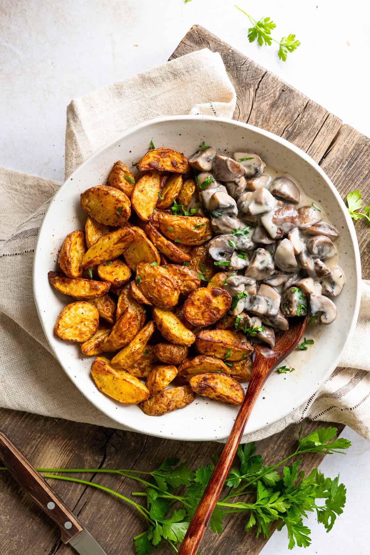 Eine helle Keramikschüssel mit einer Kartoffel-Champignon-Bowl aus der Vogelperspektive. Links liegen knusprig gewürzte, goldbraune Drillinge aus dem Airfryer, rechts oben befindet sich die cremige Champignon-Rahmsoße mit Petersilie. Ein Holzlöffel ragt nach unten rechts heraus, im Hintergrund sind rustikales Holz, ein Leinentuch und frische Kräuter zu sehen.