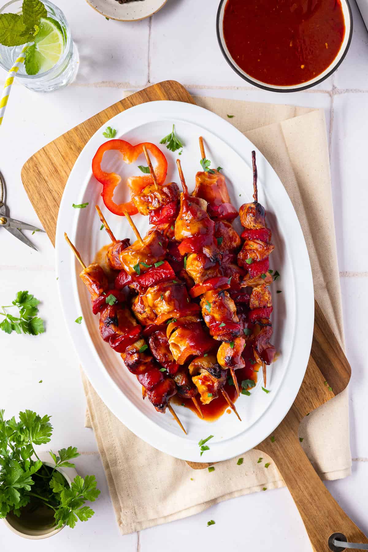 Die gegrillten Schaschlikspieße liegen glasiert mit roter Schaschliksoße auf einer ovalen Porzellanplatte und einem Holzbrett. Als Dekoration dienen frische Petersilie, eine Paprikascheibe, ein Glas Zitronenwasser mit Minze und eine extra Schale Soße auf einem hellen Fliesenspiegel.
