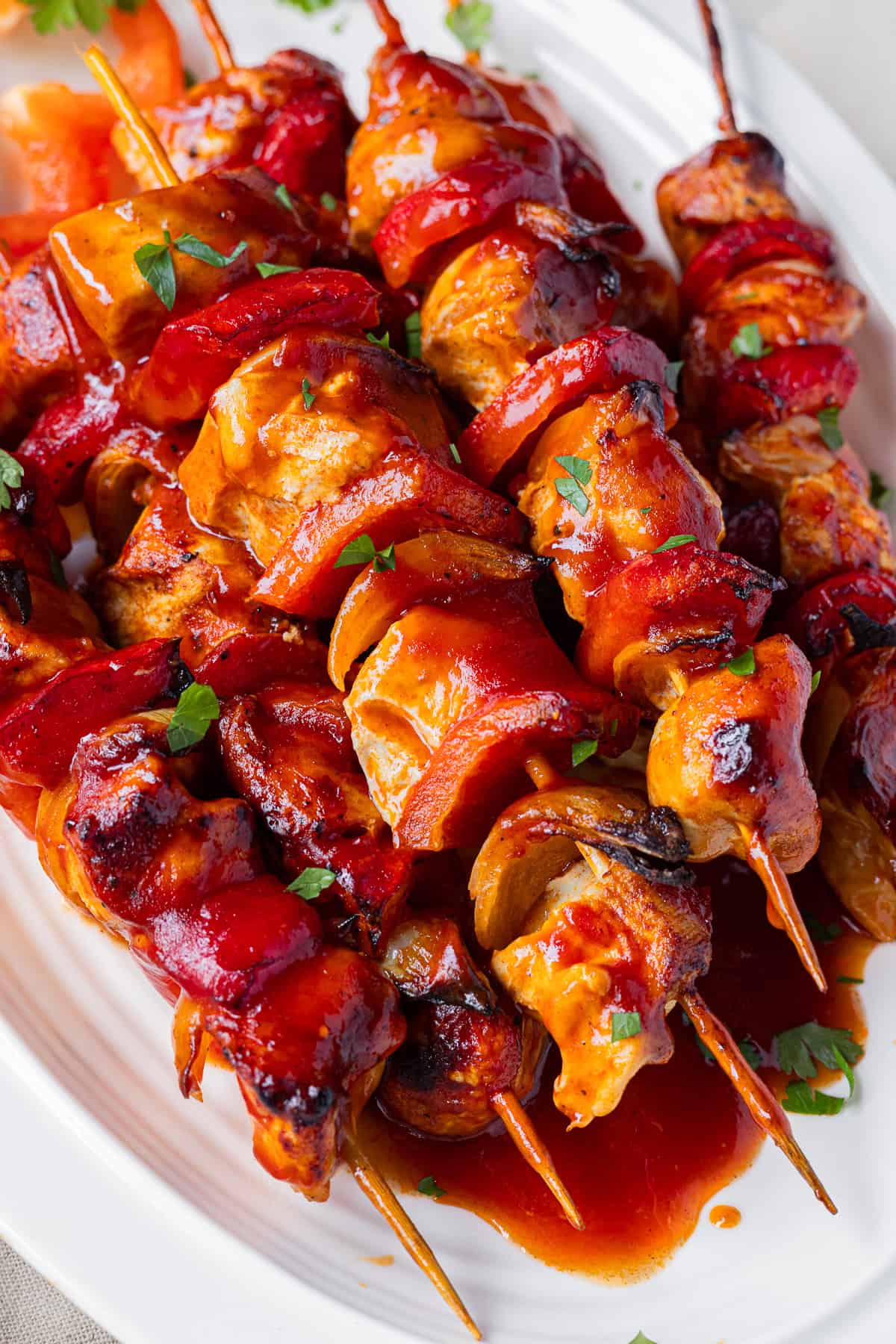 Saftiges Hähnchen-Schaschlik aus dem Airfryer, angerichtet auf einer weißen Porzellanplatte. Die gegrillten Schaschlikspieße sind dick mit einer glänzenden, roten Schaschliksoße überzogen und mit frischer Petersilie garniert. Das Fleisch und die Paprika zeigen feine Röstaromen aus der Heißluftfritteuse.