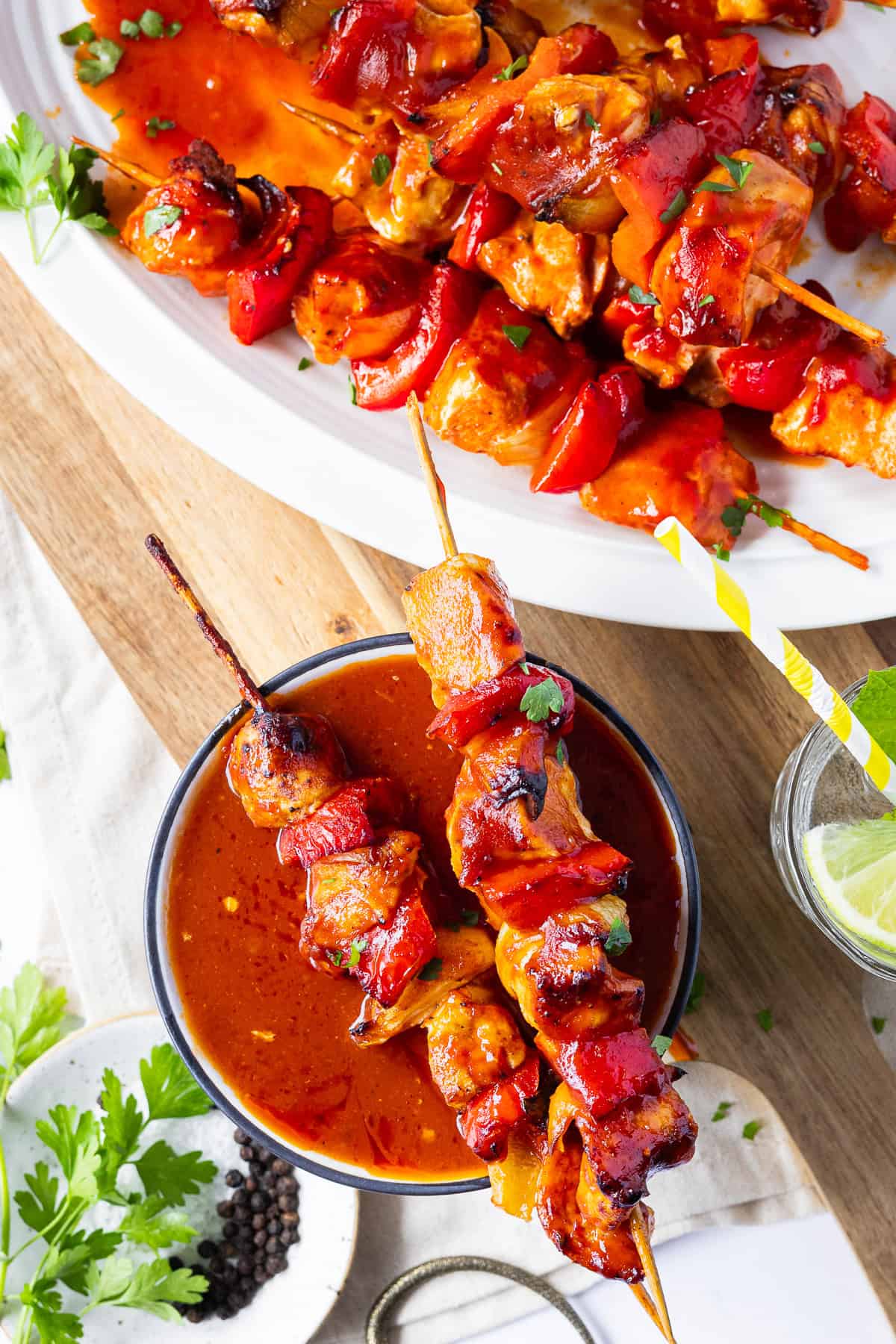 Im Vordergrund liegt ein gegrillter Schaschlikspieß aus dem Airfryer quer über einer kleinen, mit roter Schaschliksoße gefüllten Schale. Im Hintergrund sieht man eine angeschnittene Servierplatte mit weiteren Spießen, ein Holzbrett auf hellen Fliesen, Pfefferkörner und ein erfrischendes Zitronenwasser.